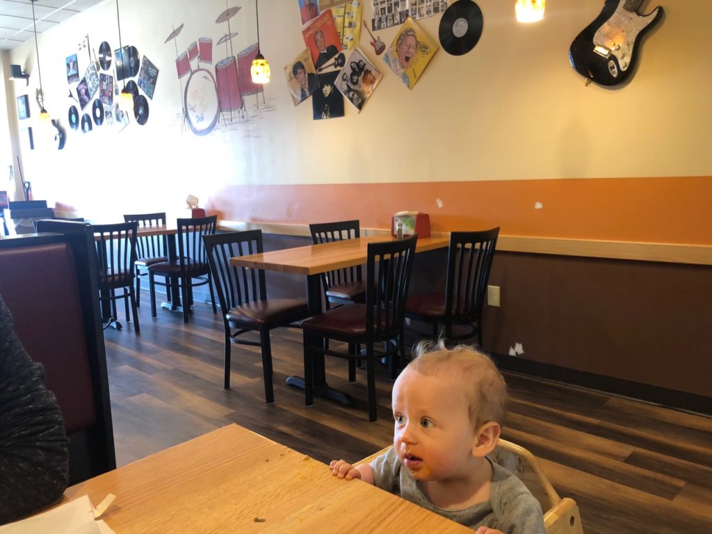 A baby sits at a cafe table with an eager look on his face
