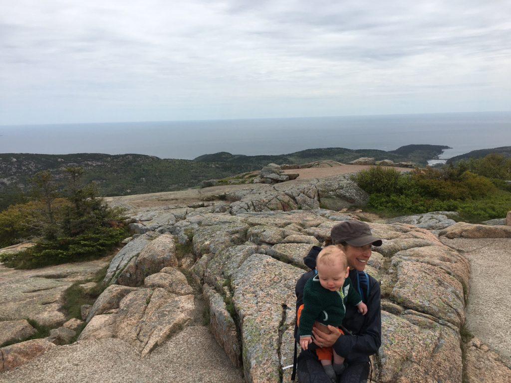 An adult holds a small child near large rocks at the top of a mountain; sea in the distance