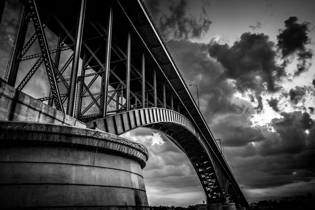Black and white image of a metal bridge seen from below. Above, there is a dramatic break in the clouds.