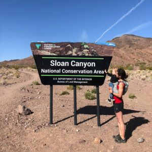 Person stands, holding a baby, in a desert landscape next to a sign that says "Sloan Canyon"