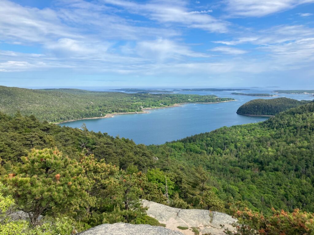 View from the top of a (modest) mountain, showing a large body of water surrounded by green, forested hills. The ocean appears in the far distance and everything is under a blue sky laced with white and grey clouds. Afternoon light.