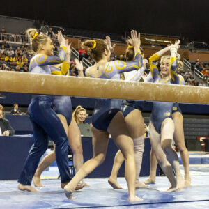 Several people in gymnastics leotards high-five each other near a balance beam