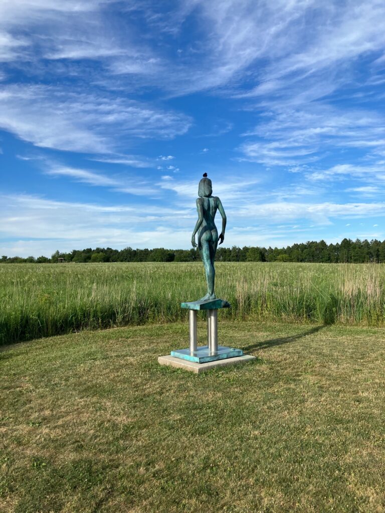 sculpture of a woman standing; there is grassland beyond her and a blue sky above