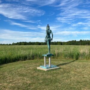 wide open green meadow under a blue sky; there's a sculpture of a naked woman in the center. There's a bird on the head of the sculpture