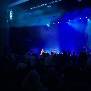 Two performers spotlighted on a stage against the silhouetted heads of a standing audience